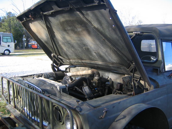 Olive drab military truck with hood open, showing engine compartment. Grille and headlights visible