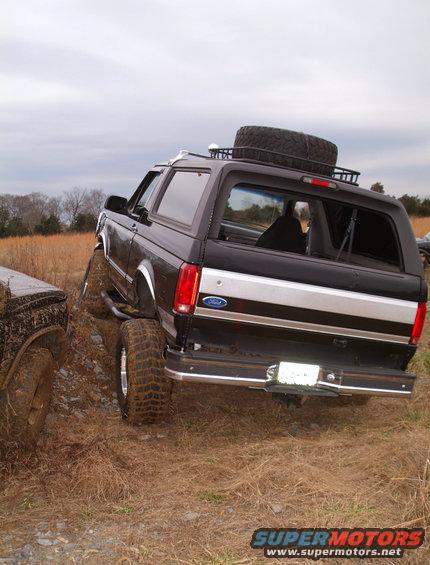 Black Ford Bronco with a spare tire on the roof rack, parked on a dirt incline next to another vehicle