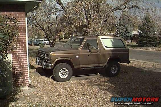 Brown Ford Bronco parked on gravel next to a brick house. Trees and a street are in the background
