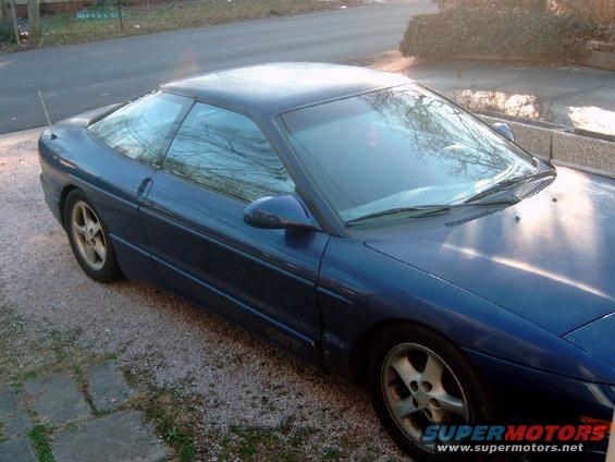 Blue coupe parked on gravel, front 3/4 view. Visible text 'SUPERMOTORS'