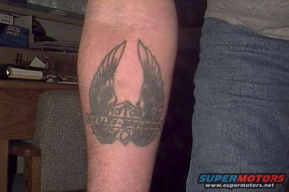 Tattoo of winged shield emblem on arm, with denim jeans and a wooden chair visible