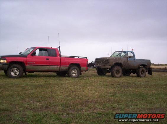 Mud-covered blue 1980s Chevrolet K5 Blazer being towed by a red Dodge Ram pickup truck on a grassy field
