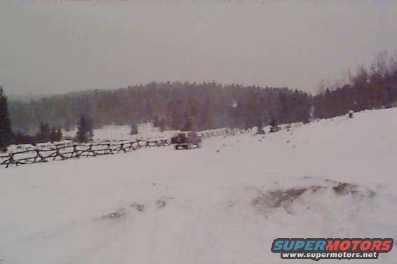 Vehicle driving on a snowy hill with a wooden fence and trees in background