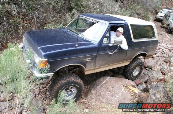 Dark blue Ford Bronco with white top, driver in white hat, navigating rocky terrain. Another SUV is visible behind it