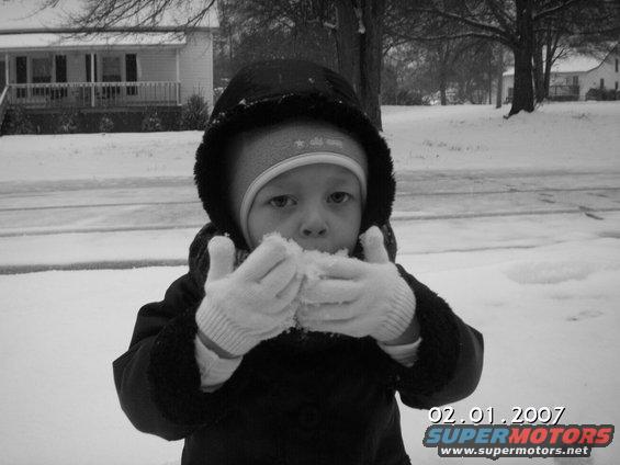 Kid in snow eating a snowball, house and trees in background