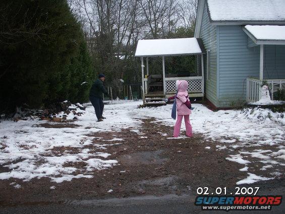 man throws a snowball at a child in a pink hooded snowsuit in a yard with snow and mud
