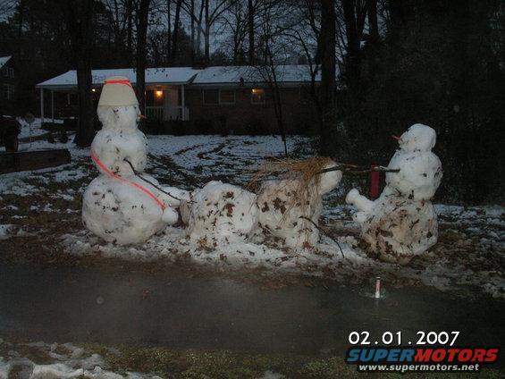 Three snowmen in a yard with a house in the background; one snowman has a bucket on its head