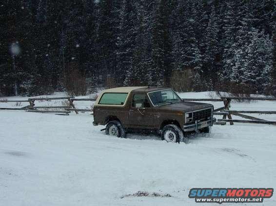 Brown Ford Bronco parked in deep snow with trees and a wooden fence in the background