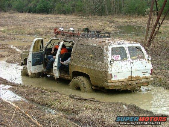 Mud-covered SUV with open doors stuck in muddy water, two people inside
