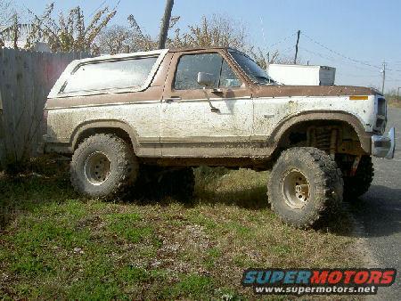 Brown and white Ford Bronco with lifted suspension and large tires, parked on grass next to a wooden fence