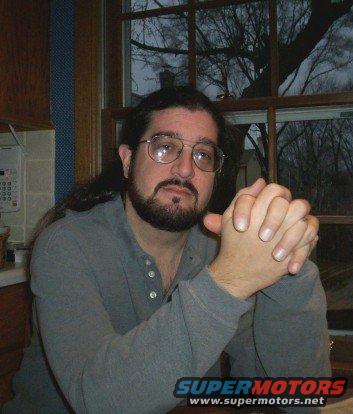 Man with beard and glasses sitting indoors, SUPERMOTORS.NET logo visible