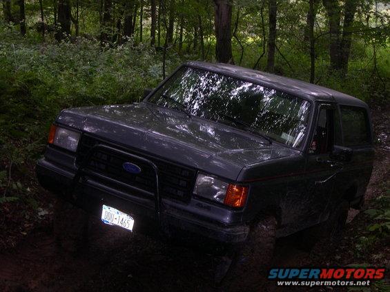 Dark gray 1987 Ford Bronco with a brush guard and off-road tires on a muddy forest trail