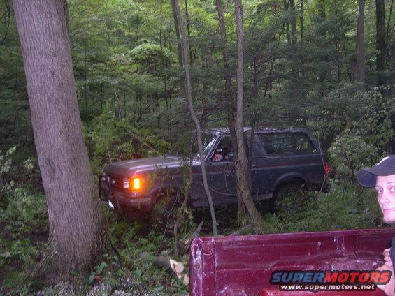 Dark Ford Bronco off-road, stuck between trees. Red pickup truck bed in foreground