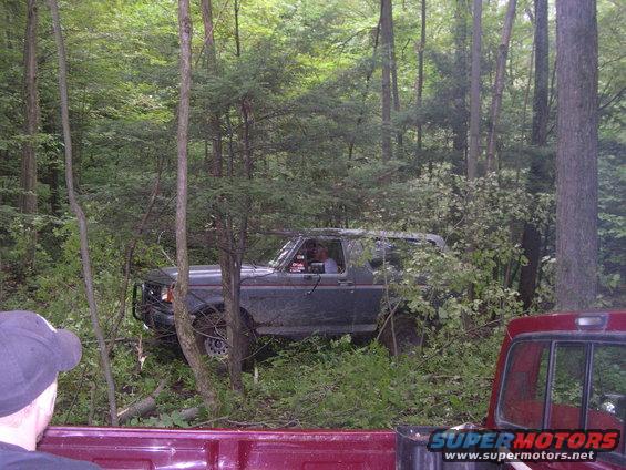 Mud-covered dark gray Ford Bronco off-road in a wooded area, front-left tire visible