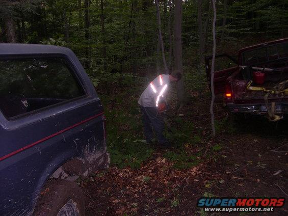 Dark blue Ford Bronco and red pickup truck in woods, guy with chainsaw working near trees