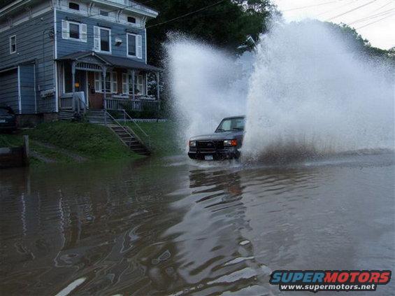 Dark SUV driving through floodwater, creating large splashes. A blue house is visible on the left
