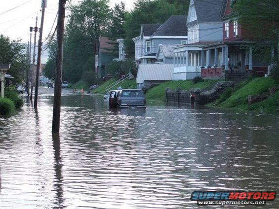 Flooded street with a dark SUV partially submerged in water. Houses line the street
