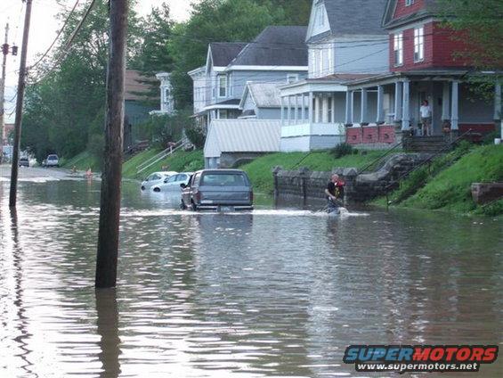 Dark SUV driving through flooded street with houses on the side