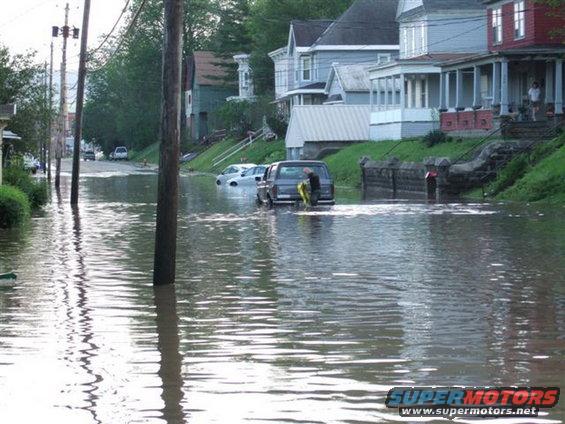 Dark van partially submerged in floodwater on a residential street, with a person standing behind it