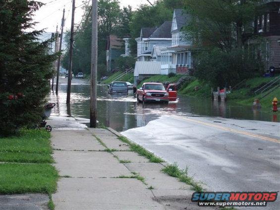 Red pickup truck with open door driving through floodwaters on a residential street
