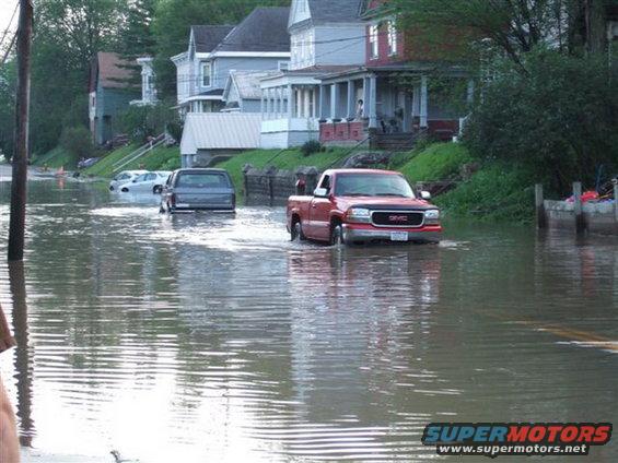 Red GMC pickup truck driving through flooded street with houses in background. Another vehicle is visible in the water
