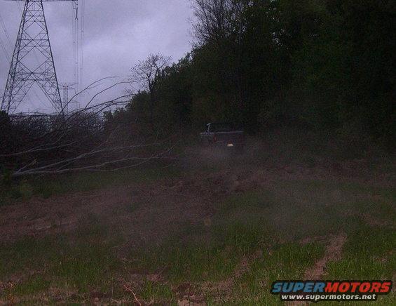 Dark 1987 Ford Bronco driving on a dirt path with trees and a power line tower in the background
