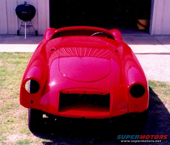 Red sports car body, no grille or headlights visible. Steering wheel is inside. Parked outside a garage with a grill nearby
