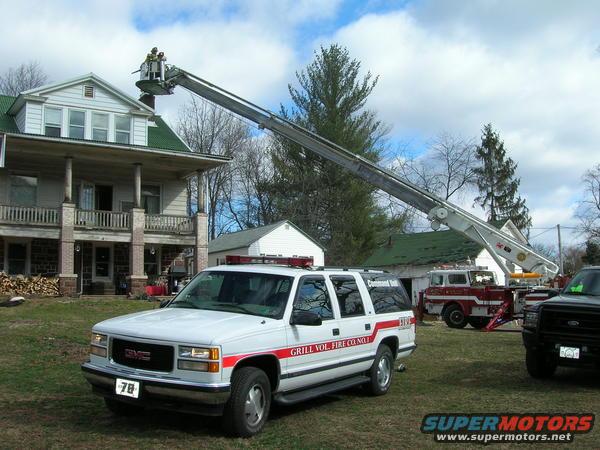 Fire truck with aerial ladder extended towards a house, GMC SUV in foreground, another fire truck behind