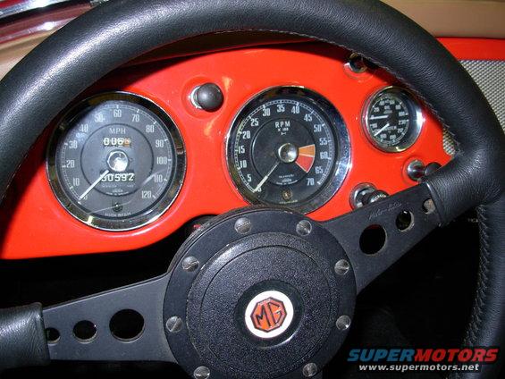 Dashboard with red housing, black steering wheel, and three gauges including speedometer and tachometer. MG logo on steering wheel