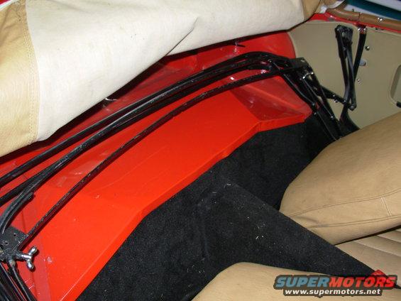 Car interior with tan seats, black carpet, and red paneling under a folded tan convertible top frame