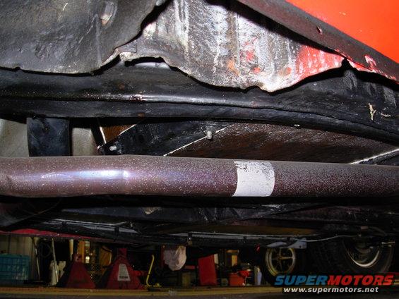 Underside view of a vehicle showing the exhaust pipe and frame components. A wheel is visible in the background