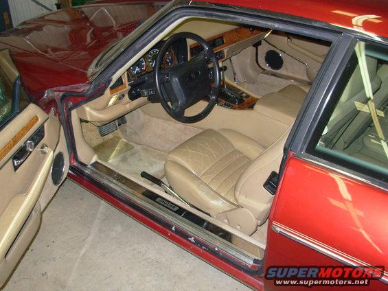 Red Jaguar XJ-S interior with tan leather seats, wood trim, and dashboard visible. The driver's door is open