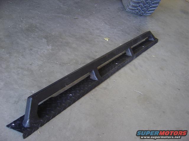 Black diamond plate step with a black metal bar, angled view, on concrete floor
