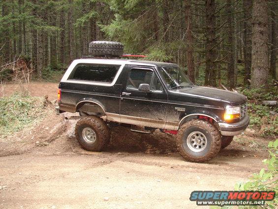 Black 1996 Ford Bronco with a spare tire on the roof, driving on a dirt trail in a forest