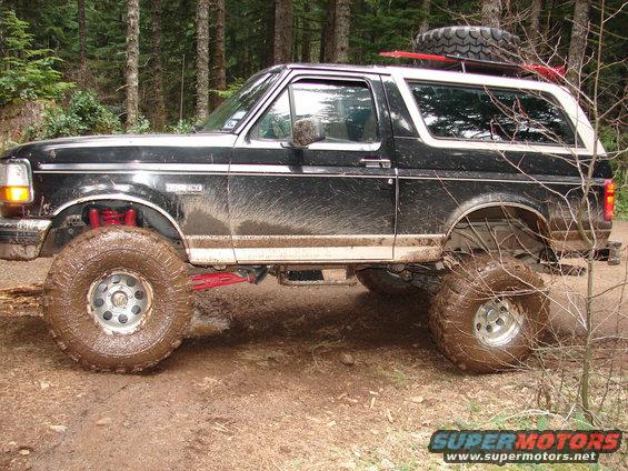 Side view of a black 1996 Ford Bronco with large mud tires, lifted suspension, and a spare tire on the roof rack, driving on a muddy trail in a forest