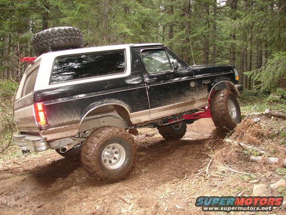 Black and white 1996 Ford Bronco with a spare tire on the roof, driving on a muddy forest trail