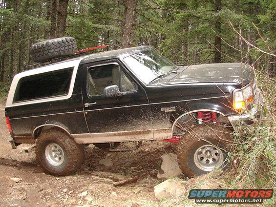 Black Ford Bronco with a spare tire on the roof, driving on a dirt trail through trees