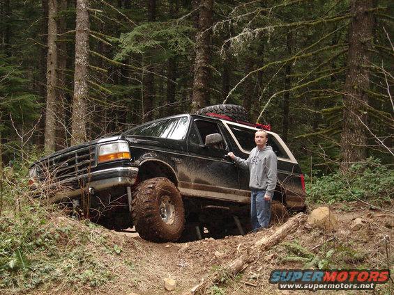 Black Ford Bronco with lifted suspension and large tires on a dirt trail, a person stands beside the driver's door