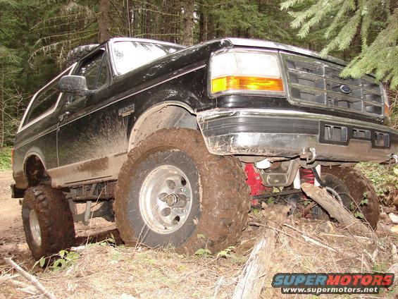 Mud-covered black 1992 Ford Bronco with lifted suspension, front-right tire stuck in dirt, in a wooded area