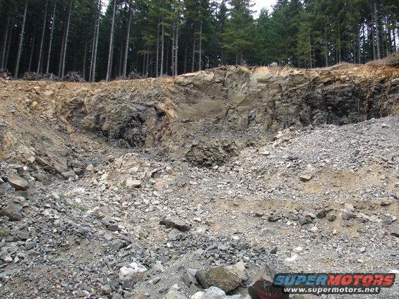 Rock quarry with steep, layered walls and a pile of rubble in the foreground, backed by a dense forest