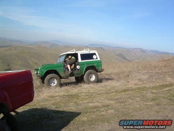 Green classic SUV with white top parked on grassy hill, person in driver's seat. Red truck in foreground