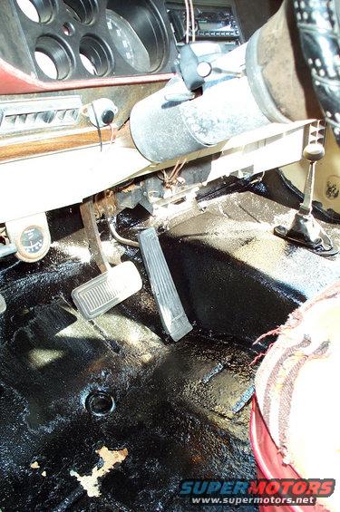 Interior view of vehicle floor with black undercoating, pedals, and dashboard visible