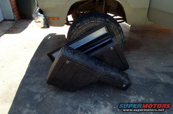 Two black plastic truck bed toolboxes stacked on concrete, with a tire and part of a vehicle in the background