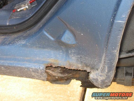 Blue vehicle rocker panel with significant rust damage and a hole exposing internal structure. A black rubber seal is visible above