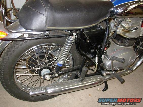 Motorcycle rear side view, showing black seat, spoked wheel, chrome exhaust, and engine details. Blue stripe on tank