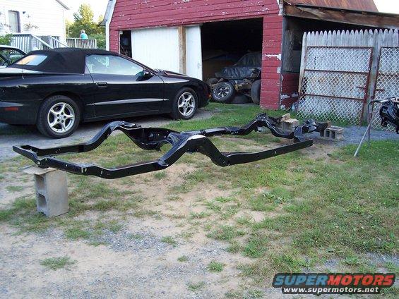 Black convertible parked next to a car frame on cinder blocks. Another vehicle is in the background inside a barn