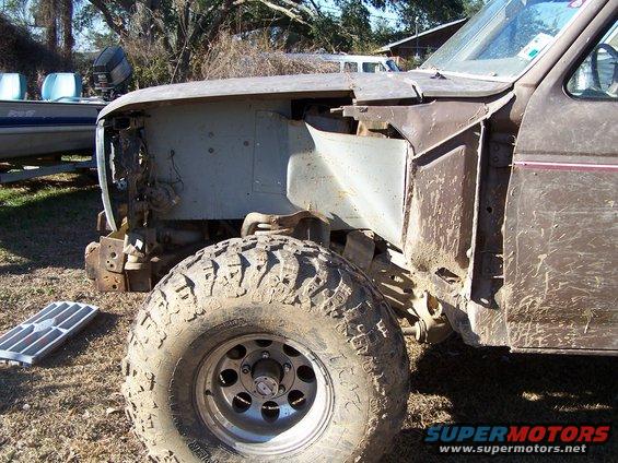 Front wheel and fender of a muddy, partially disassembled truck. A boat is visible in the background