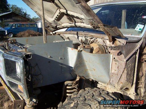 Hood open on a rusty Ford Bronco, showing the engine bay and front suspension. A blue van is parked in the background