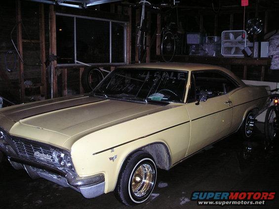 Yellow 1965 Chevrolet Impala coupe with gold wire wheels parked in a garage, 'For Sale' sign on windshield