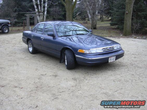 Blue 1995 Ford Crown Victoria parked on gravel, with a dark pickup truck to the left